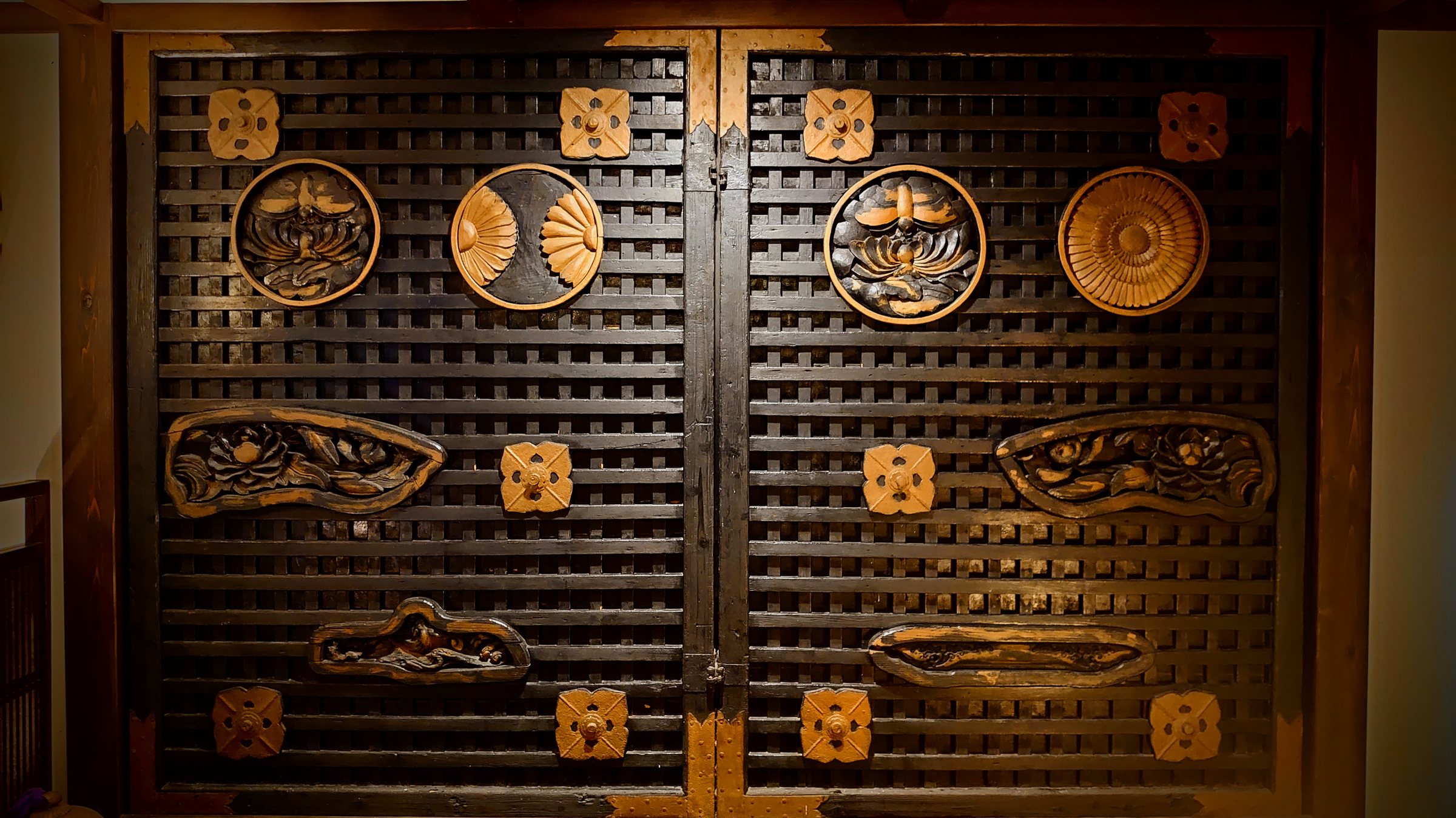 The doors of the Gomadō of Kunōzan Tōshōgū, now standing in the lobby at Shirakabe: dark timber grillwork inset with gilded roundels of lotus, chrysanthemum and fan