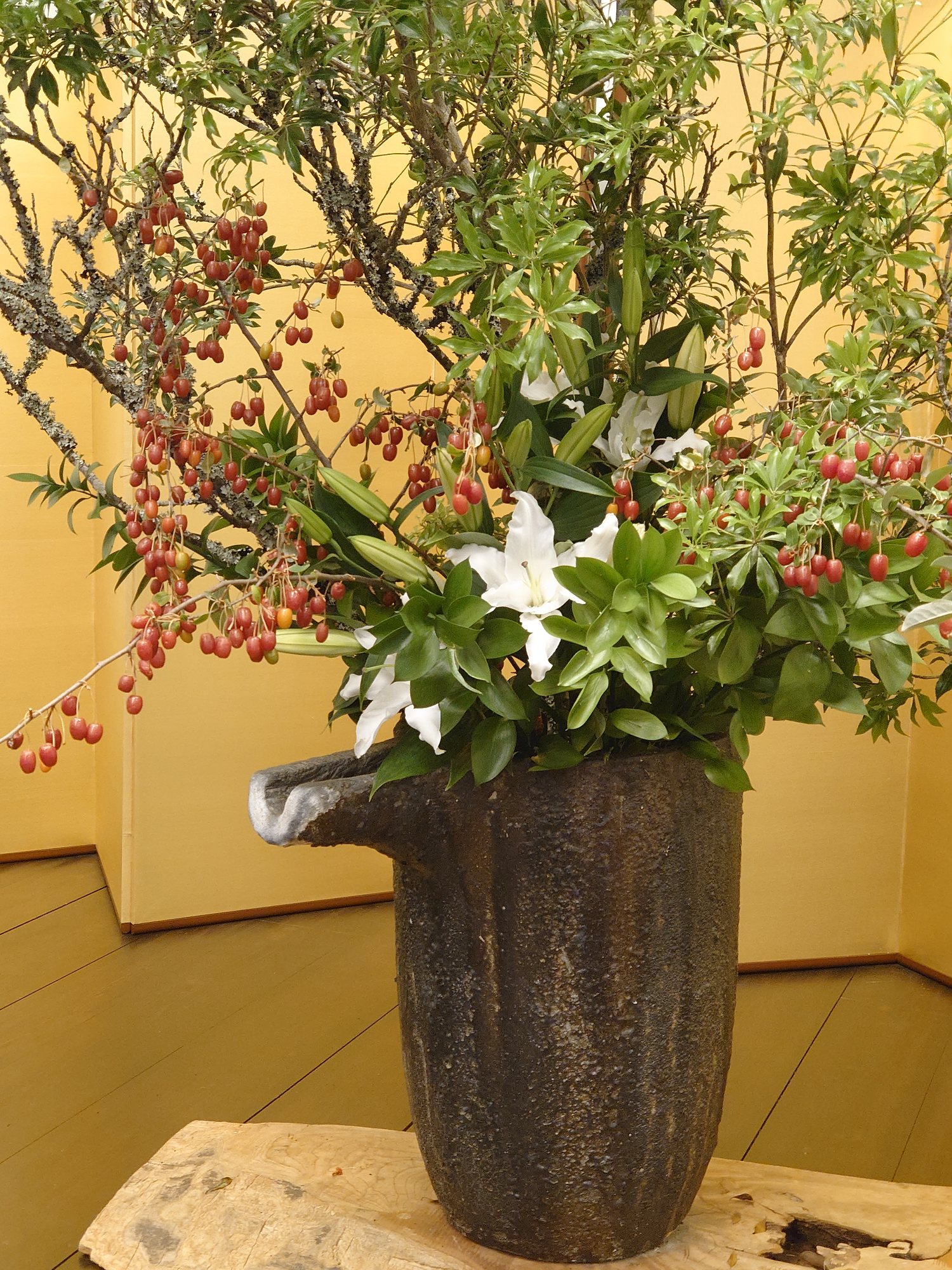 White lilies and berried gumi branches arranged in a rough stone vessel, set against a gold folding screen